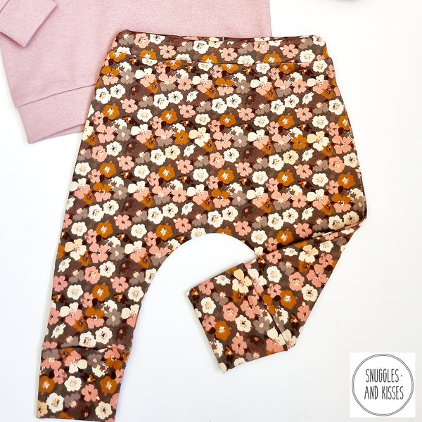 photo is of a pair of baby leggings in a small scale floral print. The background colour of the print is a warm brown colour and the flowers, which are like Mallow flowers, are cream, brown and pink