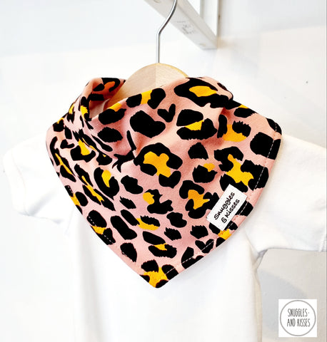 pink leopard print dribble bib with Snuggles and Kisses label positioned to the left hand side as worn. Shown on a hanger, worn with a plain white baby vest. The leopard print has a pink background with a black and yellow leopard spot.